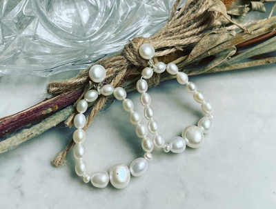 A pair of white freshwater pearl earrings with silver hooks, displayed on a twine with a natural leaf background.